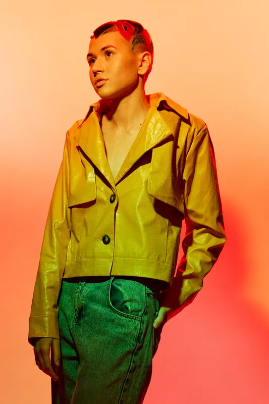 Young woman with short hair wearing a yellow jacket posing against an orange background with dramatic lighting and modern fashion style.
