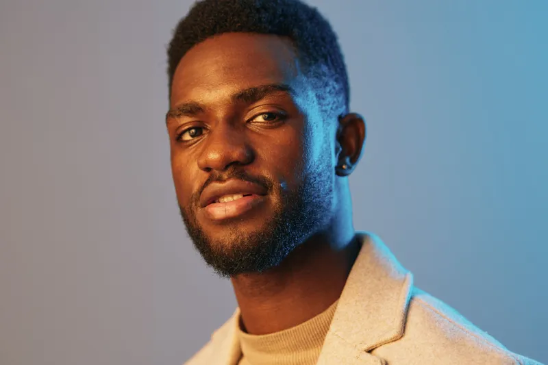 Confident young African man in stylish cream coat smiles against a vibrant blue background, showcasing modern fashion and self assurance