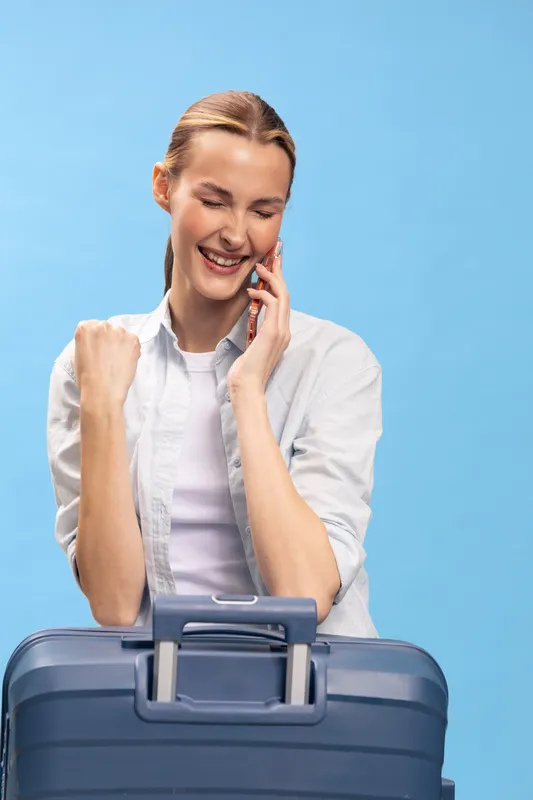 Happy woman with suitcase talking on phone, smiling and celebrating with fist pump. Travel concept, excitement, positive emotions, and successful vacation preparation on blue background.