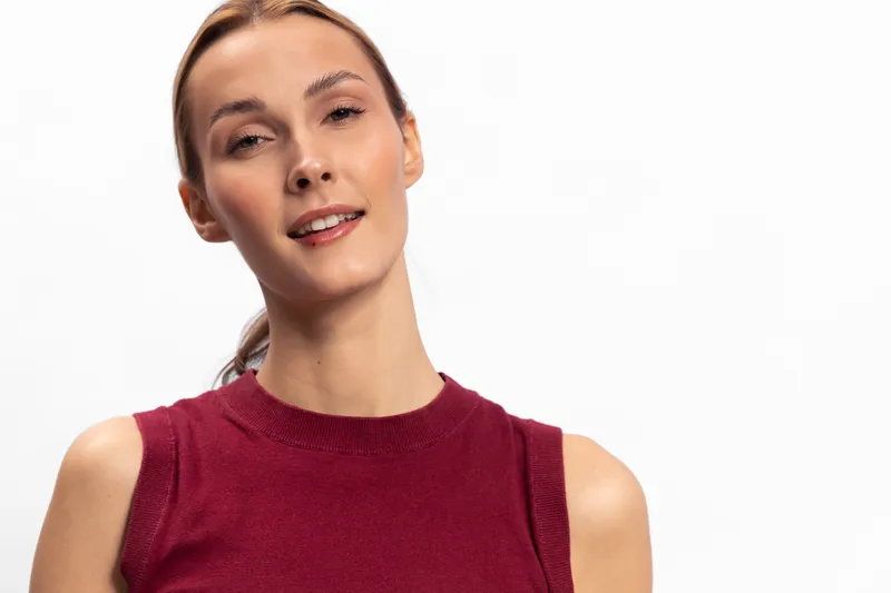 Smiling young woman in sleeveless maroon top stands against white background. She looks relaxed and confident with natural makeup and smooth skin in bright studio lighting.