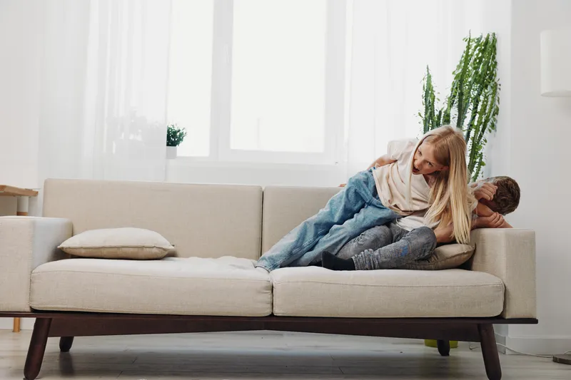 Playful siblings laughing on beige sofa in bright living room with large windows and green plants. Concept for family fun, childhood joy, and home comfort.
