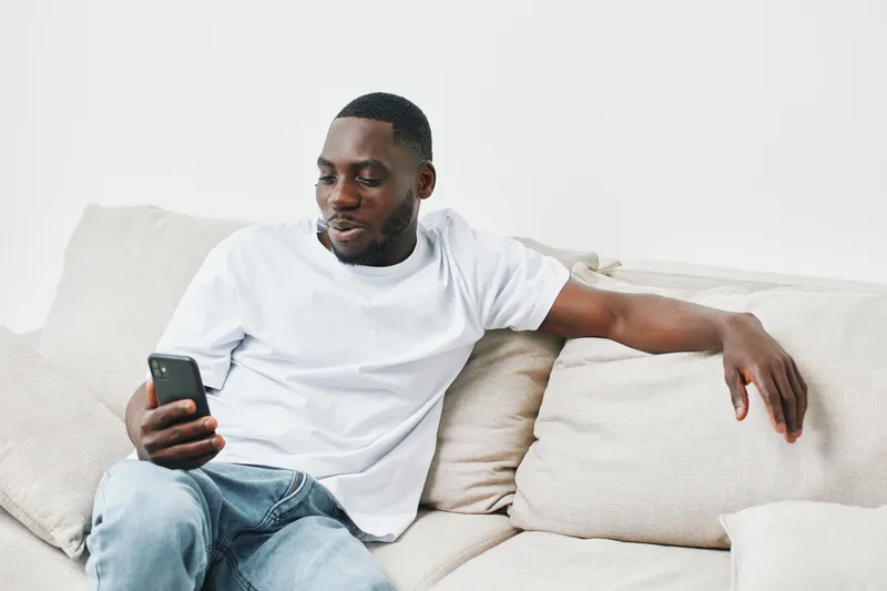 Young Black man in casual attire enjoying his phone on a cozy couch, showcasing a relaxed lifestyle and modern technology interaction