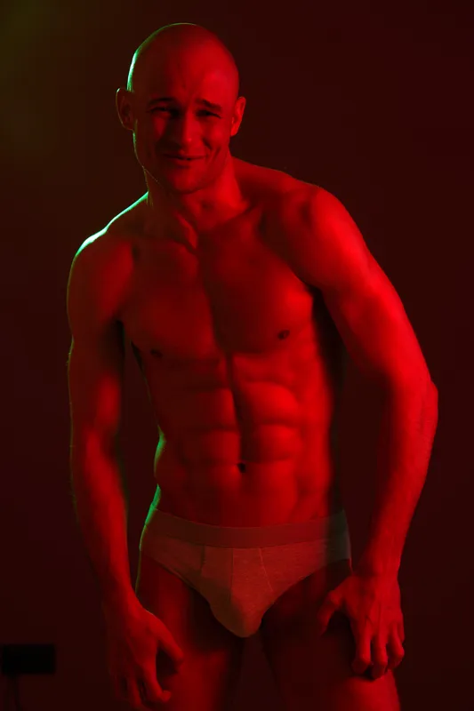 Fit man posing under dramatic lighting, showcasing a muscular physique with intense shadows and highlights, highlighting strength and determination in a striking manner.