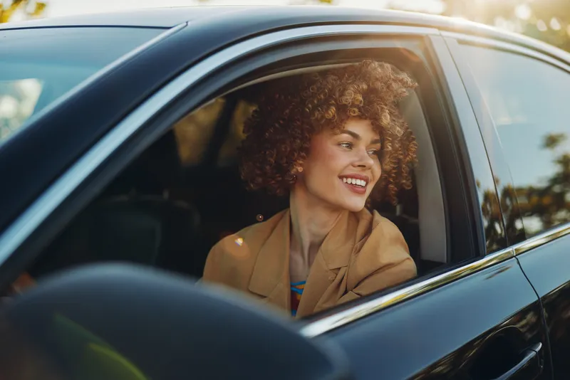 Smiling woman sits in a car, wearing a tan blazer and curly hair, captured with retro film tones.