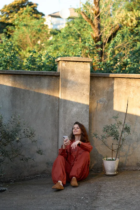 Woman lifestyle outdoor scene shows a relaxed moment with a woman sitting by a wall, wearing red outfit and using a smartphone, calm casual mood