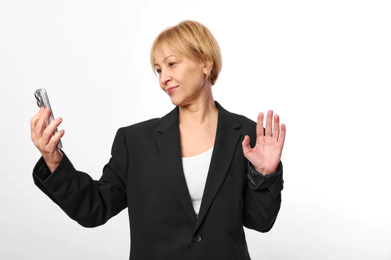 Woman business blazer smartphone skeptical gesture communication isolated portrait, blond woman raises hands holding mobile device, unsure expression on plain white background.
