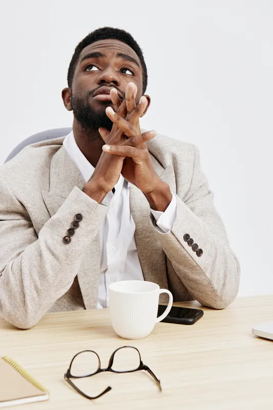 Thoughtful man with dark skin, beard, and short hair sitting at desk in beige suit and white shirt, looking upward with contemplative expression, hands clasped near face, on plain white background, st