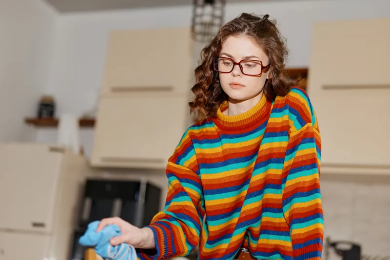 Young woman with curly hair and glasses tidies a bright kitchen, wearing a colorful striped sweater and focused expression as she folds, preparing the space