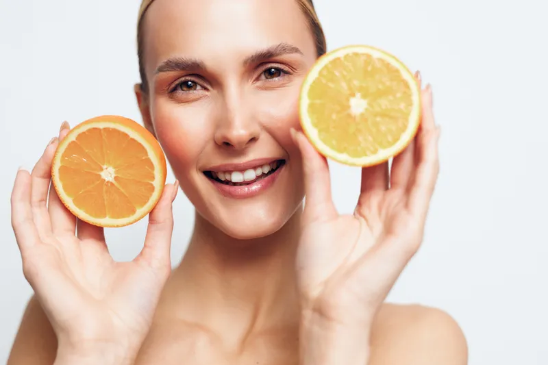 Happy young woman holds two halves of fresh orange fruit near face smiles with healthy glowing skin isolated on white background natural beauty concept.