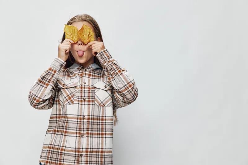 Child girl in checkered shirt holding autumn leaves over eyes and sticking tongue out for playful expression on light background with fun mood and seasonal concept.