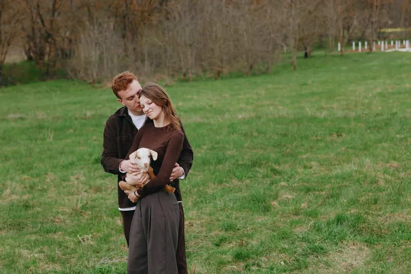 Young couple embraces tenderly in a green field while the woman holds a small white goat, sharing a peaceful moment of connection and warmth in nature.