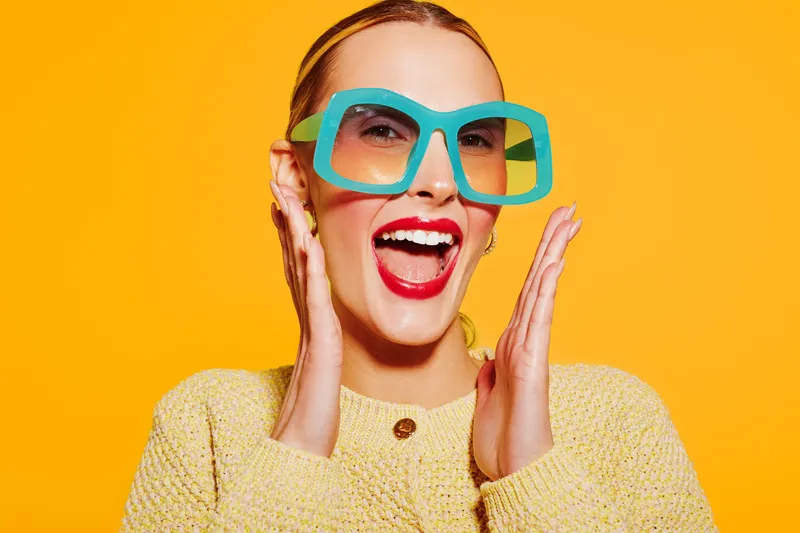 Happy young woman with bright red lipstick and large blue sunglasses smiles widely on vibrant orange background. Concept of joy, fashion, summer, and positive emotions.