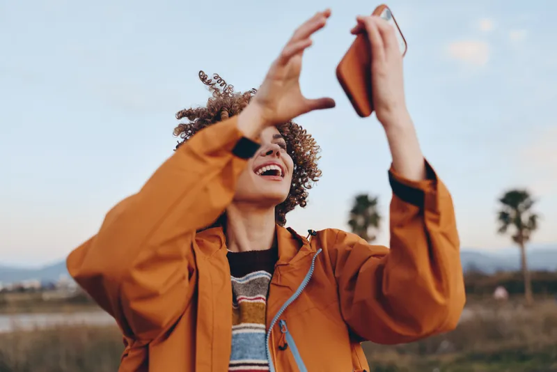 Woman lifestyle outdoors, laughing and holding a smartphone for a selfie, orange jacket with hood, curly hair lifted by a breeze, sunlit field background, relaxed pose