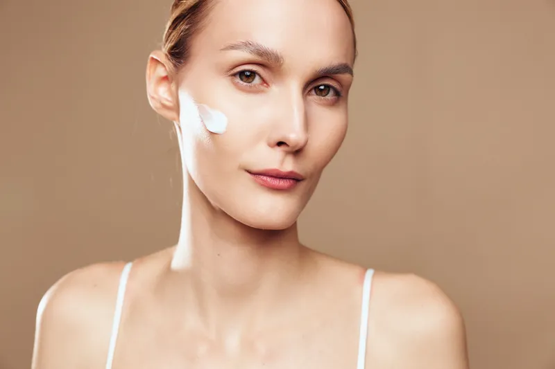 Young woman with natural skin applies moisturizing cream on face looking at camera with soft expression isolated on beige background, skincare concept and beauty routine.