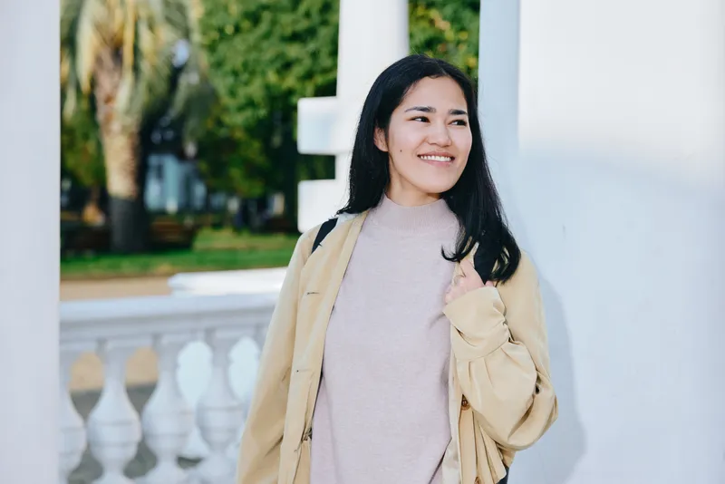 Woman smiling outdoors in a light beige coat and soft pink sweater, standing near a white balustrade with greenery behind, conveying casual fashion and cheerful mood.