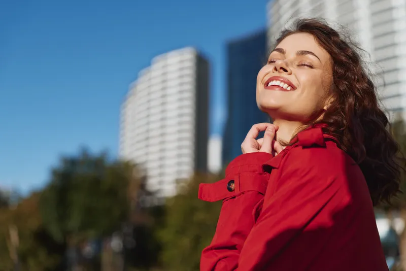 Smiling woman in a red coat outdoors, basking in sun with a city backdrop. Concept of confidence, fashion, and well being. Soft daylight creates a warm, uplifting mood.