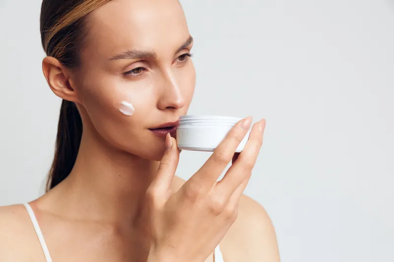 Young woman with clear skin applying moisturizer on face holding white cream jar, soft natural light, minimal makeup, smooth skin texture, skincare routine concept