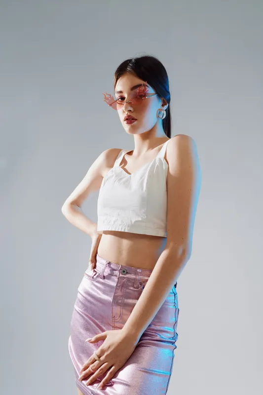 Young woman in a white crop top and shiny pink skirt poses confidently with pink sunglasses and earrings. Studio shot with soft lighting. Concept of woman beauty fashion.
