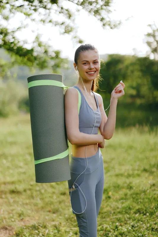 Young woman in athletic wear smiling while holding yoga mat outdoors, embodying a healthy lifestyle in nature