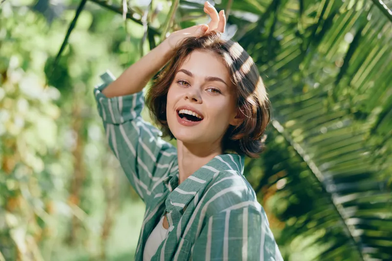 Cheerful woman enjoying warm sunlight in a lush garden, smiling with carefree energy among tropical leaves and soft greenery.