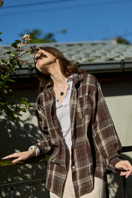 Relaxed woman outdoors wearing a plaid shirt over a white top, gazing at blossoms. Natural light highlights calm mood, casual style, and connection with nature