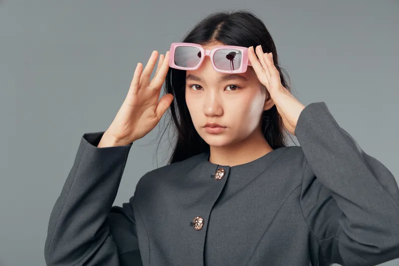 Young woman wearing oversized pink sunglasses and gray jacket with decorative buttons on gray background looking at camera with serious expression and long black hair.