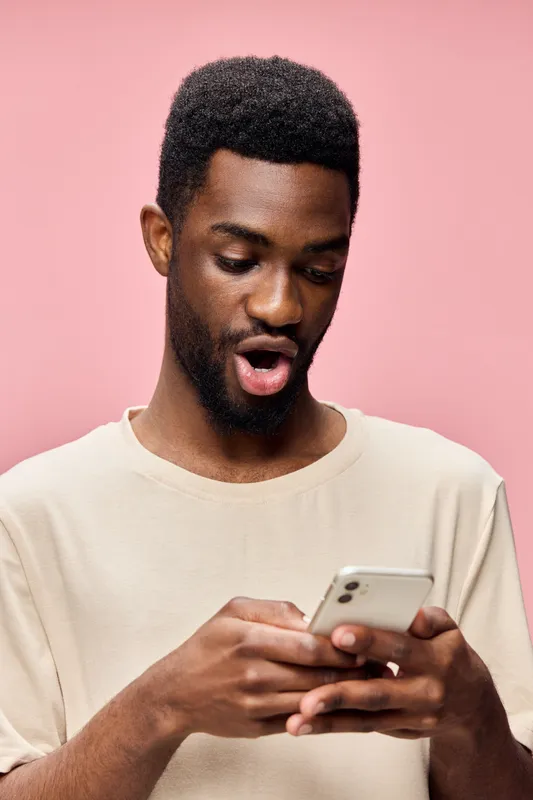 Young African man expressing surprise while using a smartphone against a pink background, casual clothing, and vibrant colors creating a modern digital lifestyle mood