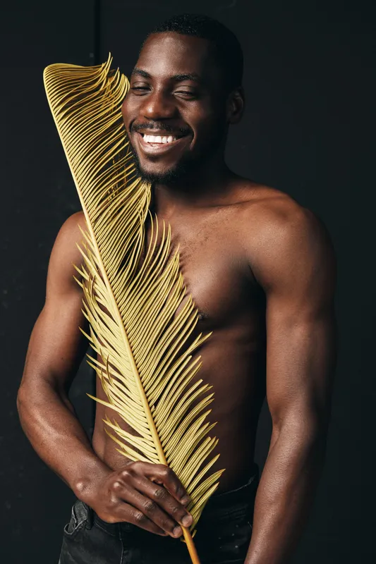 African man with dark skin holding large yellow palm leaf in creative conceptual style, smiling with closed eyes, bare chest, isolated on black background for visual art series.