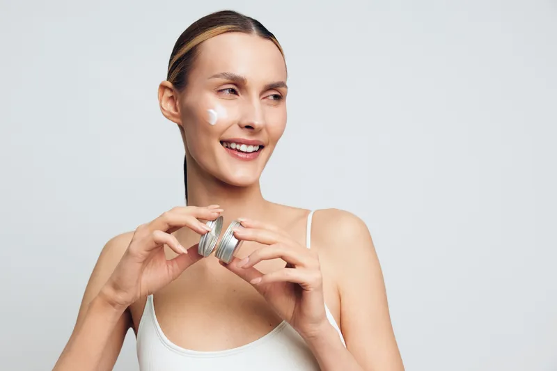 Smiling woman applying white face cream on cheek holding jar. Close-up on light background. Concept of skincare, beauty routine, self-care, and natural wellness. Soft lighting.