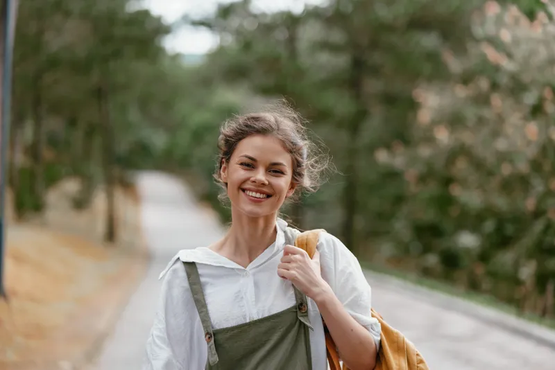 Young woman walking along a forest path, wearing casual clothing, smiling joyfully with a backpack, surrounded by greenery, evoking a sense of adventure and happiness