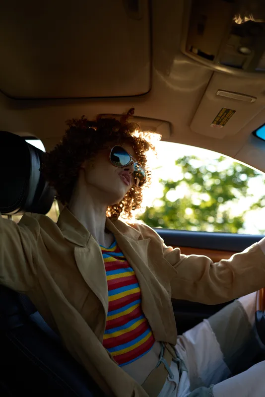 Woman driving a car, casual retro style, striped shirt and sunglasses, sunlit interior, relaxed posture, warm tones, candid mood, outdoors greenery in motion