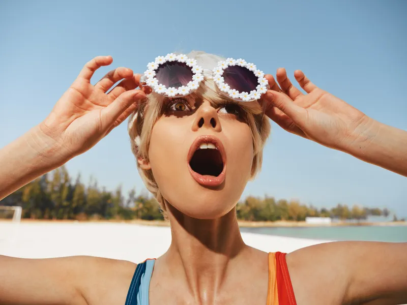 A joyful young woman at the beach wears floral sunglasses, eyes wide, mouth open in surprise, capturing a carefree moment under a bright blue sky.
