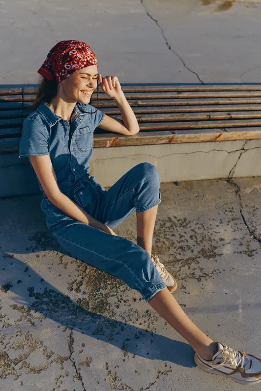 lifestyle woman in modern boho-western denim street style sitting outdoors with a red bandana and casual sneakers in warm film color tones