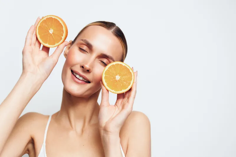Smiling woman holding orange slices near her cheeks, healthy citrus skincare concept, fresh vitamin C feeling, soft studio light, clean white background, youthful beauty portrait