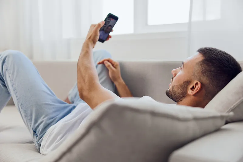 Relaxed man enjoying a video call at home, wearing casual attire, showcasing comfort and modern lifestyle in a bright living room setting.