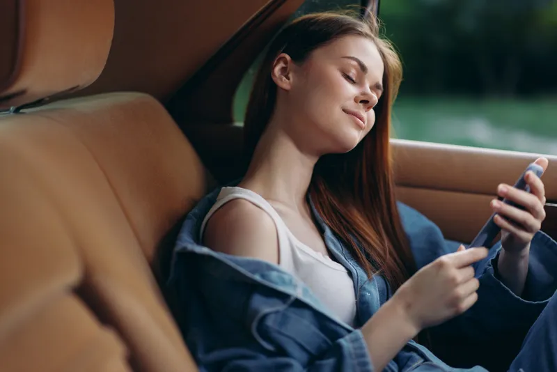 Young woman with long hair relaxing in a car while using her smartphone, wearing casual clothes, enjoying a serene moment in nature.