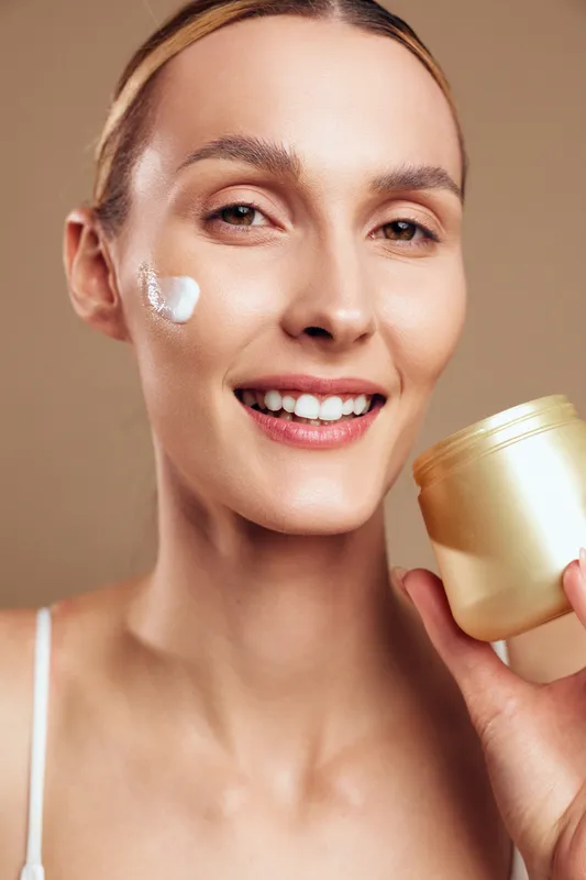 Smiling young woman holding jar of cream applying moisturizer on face with smooth skin natural beauty isolated on beige background skincare concept.