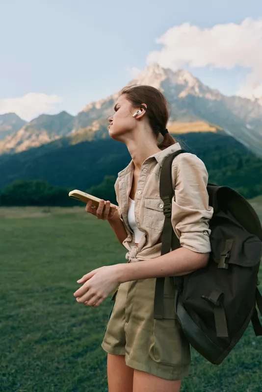 Young woman traveler with backpack holds smartphone looks away thoughtful stands on green field with mountain landscape background during outdoor adventure lifestyle.