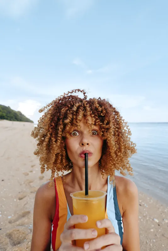 Young woman with curly hair on a sandy beach drinks orange juice through a straw, enjoying a sunny day by the shore and a refreshing moment, framed against a clear blue sky and calm water.