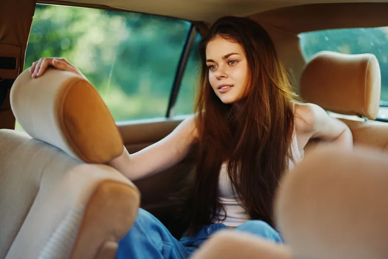 Young woman with long hair sitting in the backseat of a car, reflecting on life while enjoying the serene green background during a sunny day.