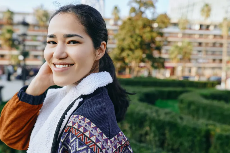 Woman with a warm smile wearing a patterned jacket and scarf outdoors in a sunny park, casual fashion, natural light, candid mood, everyday style and outdoor charm