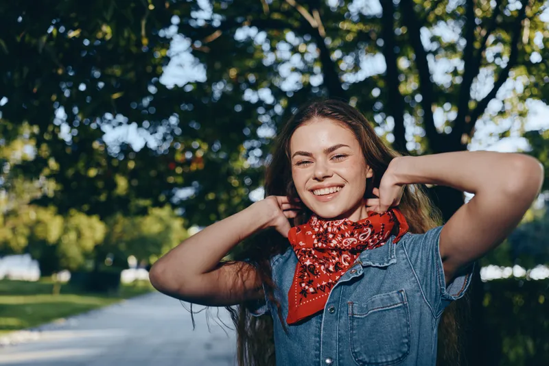 Lifestyle woman in modern boho-western denim street style wearing a red bandana, smiling outdoors under dappled sunlight with film color tones and relaxed mood.