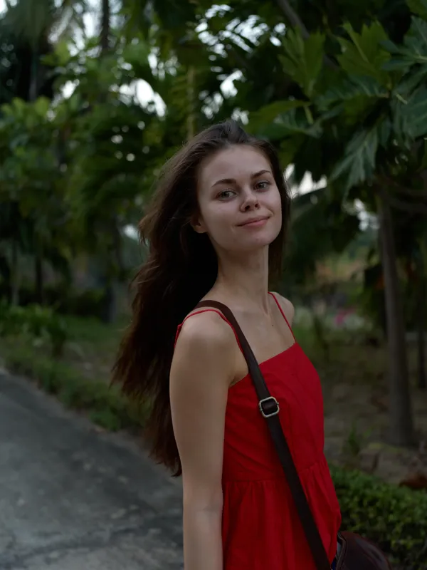 young woman wearing red dress walking outdoors in natural green environment, smiling softly with long hair flowing, casual summer style portrait