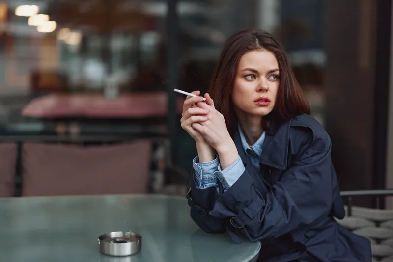 A young woman with long brunette hair sits at a glass table in a city cafe, smoking a cigarette and looking thoughtful.