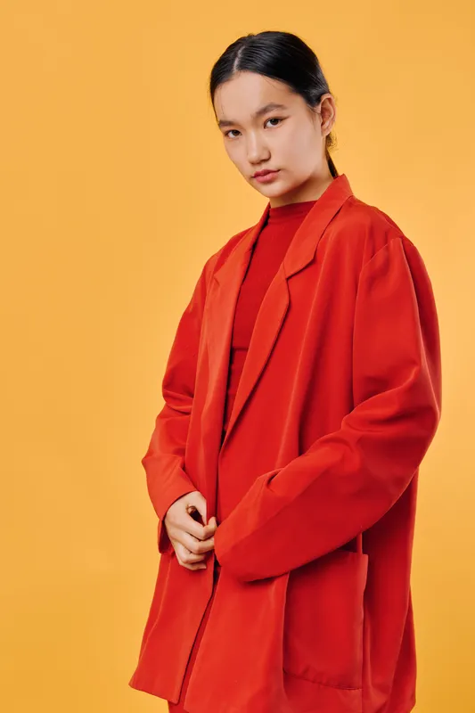 Young woman wearing oversized red blazer and red top posing against orange background. Female model with dark hair tied back, serious expression, fashion style and studio portrait.