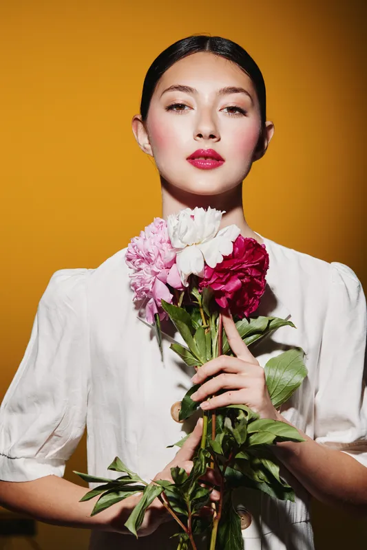 Fashionable young woman with bright makeup holds mixed peonies bouquet. Studio portrait on orange background. Concept for beauty, creative, and fashion editorial themes.