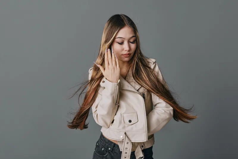 Young woman with long hair wearing beige jacket posing with closed eyes and hand near face on gray background. Fashion model with casual style and confident expression.
