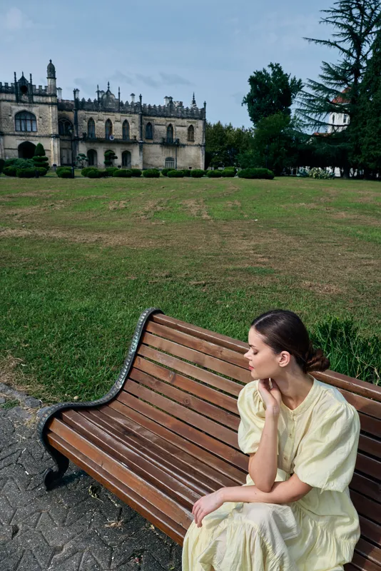 A woman in a light dress sits on a wooden bench in a park, looking thoughtful as a historic castle rises behind and green grounds extend, creating a calm outdoor landscape.