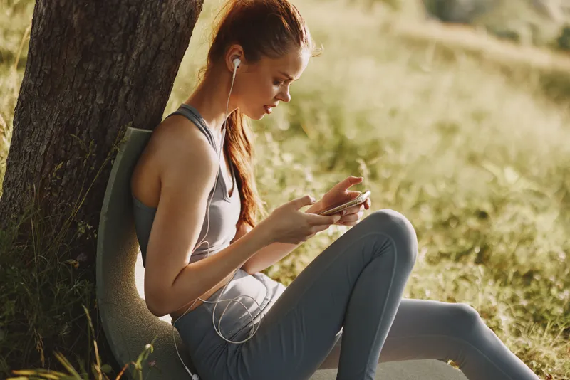 Young woman in activewear enjoying music on her smartphone while sitting outdoors, surrounded by greenery and sunlight, evoking a sense of relaxation and wellness