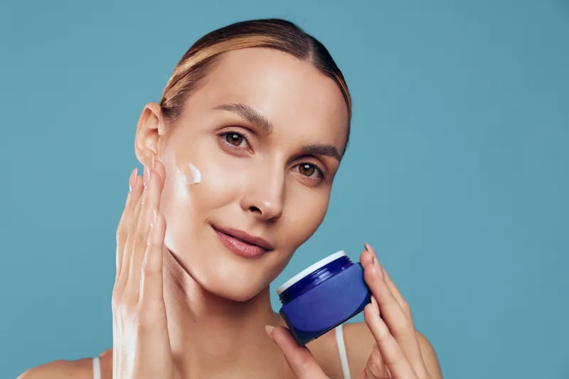 Young woman applies moisturizing cream on her face holds jar of skincare product looks at camera with soft natural makeup isolated on blue background beauty and health concept